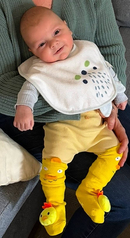 Milo a rejoint le concours — aidez-le/la à gagner de superbes lots ! adult_hand, baby, bib, child, clothing, comfort, couch, cute, duck_design, face, green_sweater, head, home, indoor, infant, person, relaxed, sitting, smiling, yellow_pants