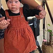 Mya participe au concours pour gagner de l'argent avec cette photo : baby, toddler, child, headband, bow, corduroy_dress, orange_dress, black_top, gold_shoes, holding_hands, adult_hand, lamp, floor_lamp, potted_plant, stroller, bracelet, glass_table, indoor, portrait, serious_face