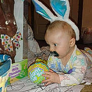 Jaxson is registered to the contest to win money with this photo: baby, bunny_ears, mustache, lollipop, chocolate_bunny, blanket, indoor, child, playful, colorful, toy, floor, door, carpet, pajamas, patterned_clothing, cute, holiday, easter, curious