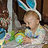 baby, bunny_ears, mustache, lollipop, chocolate_bunny, blanket, indoor, child, playful, colorful, toy, floor, door, carpet, pajamas, patterned_clothing, cute, holiday, easter, curious