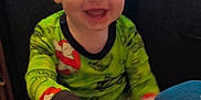 Austin is registered to the contest to win money with this photo: toddler, child, smiling, pajamas, green_clothing, hand, indoor, television, furniture, happy, face, person, young_child, couch, living_room, hair, sitting, bright_colors, playful, home