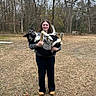 Stormy is registered to the contest to win money with this photo: woman, dog, outdoor, forest, trees, grass, smile, holding, german_shepherd, pet, nature, casual_clothing, autumn, bare_trees, animal, person, happy, daylight, ground, leaves