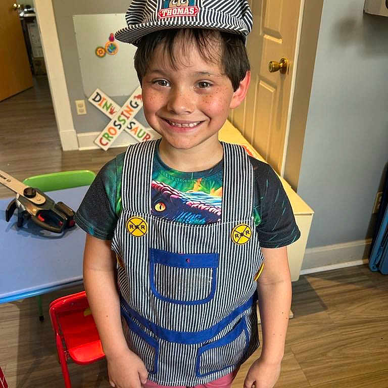 Raffael is registered to the contest to win money with this photo: boy, casual_clothing, chair, child, door, happy, hat, indoor, light_switch, overalls, person, playroom, portrait, shorts, smiling, table, toy, tshirt, wall_art, wooden_floor