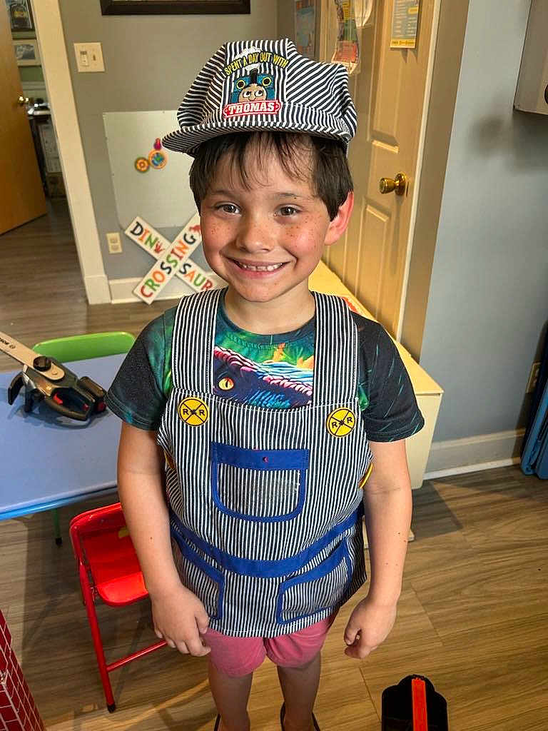 Raffael is registered to the contest to win money with this photo: child, smiling, hat, overalls, shorts, tshirt, indoor, wooden_floor, toy, table, chair, door, light_switch, wall_art, person, playroom, happy, boy, casual_clothing, portrait