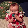 baby, child, pacifier, floral_dress, smiling, toy, cow, bed, pillow, fabric, hand, face, person, indoors, cute, playful, red, green, pink, clothing