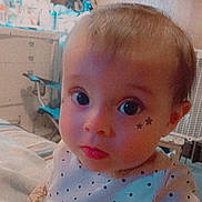 Oakley is registered to the contest to win money with this photo: baby, infant, child, portrait, close_up, wide_eyes, star_stickers, polka_dot_clothing, bib, indoor, bedroom, home, dresser, fan, blanket, seated, looking_at_camera, candid, colorful_lighting, cute