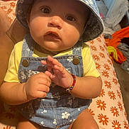 Eliana is registered to the contest to win money with this photo: baby, child, hat, denim, overalls, bracelet, hand, bed, blanket, pattern, flower, orange, yellow, skin, person, indoor, clothing, cute, young, portrait