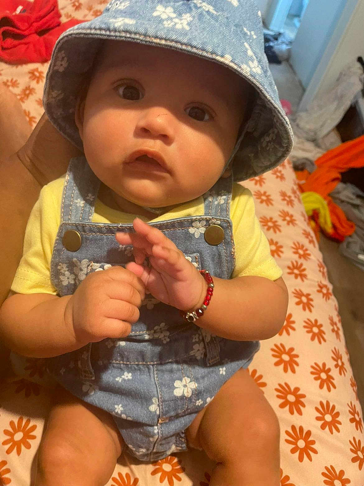 Eliana is registered to the contest to win money with this photo: baby, child, hat, denim, overalls, bracelet, hand, bed, blanket, pattern, flower, orange, yellow, skin, person, indoor, clothing, cute, young, portrait