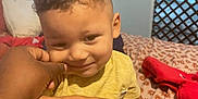 Kason is registered to the contest to win money with this photo: child, toddler, yellow_shirt, bedroom, bed, pillow, hand, smile, curly_hair, person, indoor, cozy, patterned_bedspread, cute, playful, skin, face, cheek, casual_clothing, furniture
