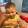child, toddler, yellow_shirt, bedroom, bed, pillow, hand, smile, curly_hair, person, indoor, cozy, patterned_bedspread, cute, playful, skin, face, cheek, casual_clothing, furniture