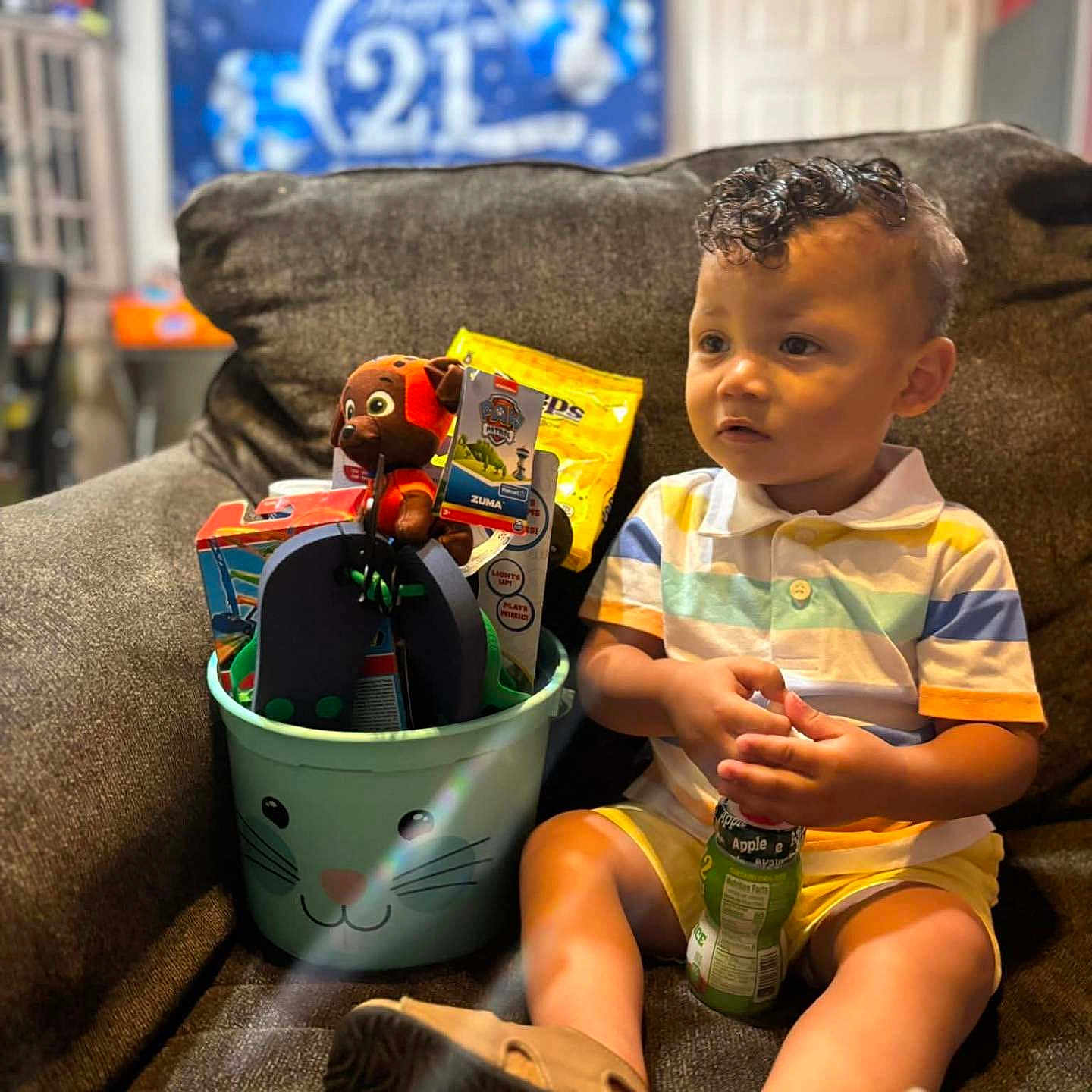 Kason is registered to the contest to win money with this photo: banner, brown_couch, bucket, celebration, child, couch, curly_hair, flip_flops, footwear, happy_21, home, indoor, juice_box, person, plush_toy, smiling_face, striped_shirt, toddler, toys, yellow_shorts