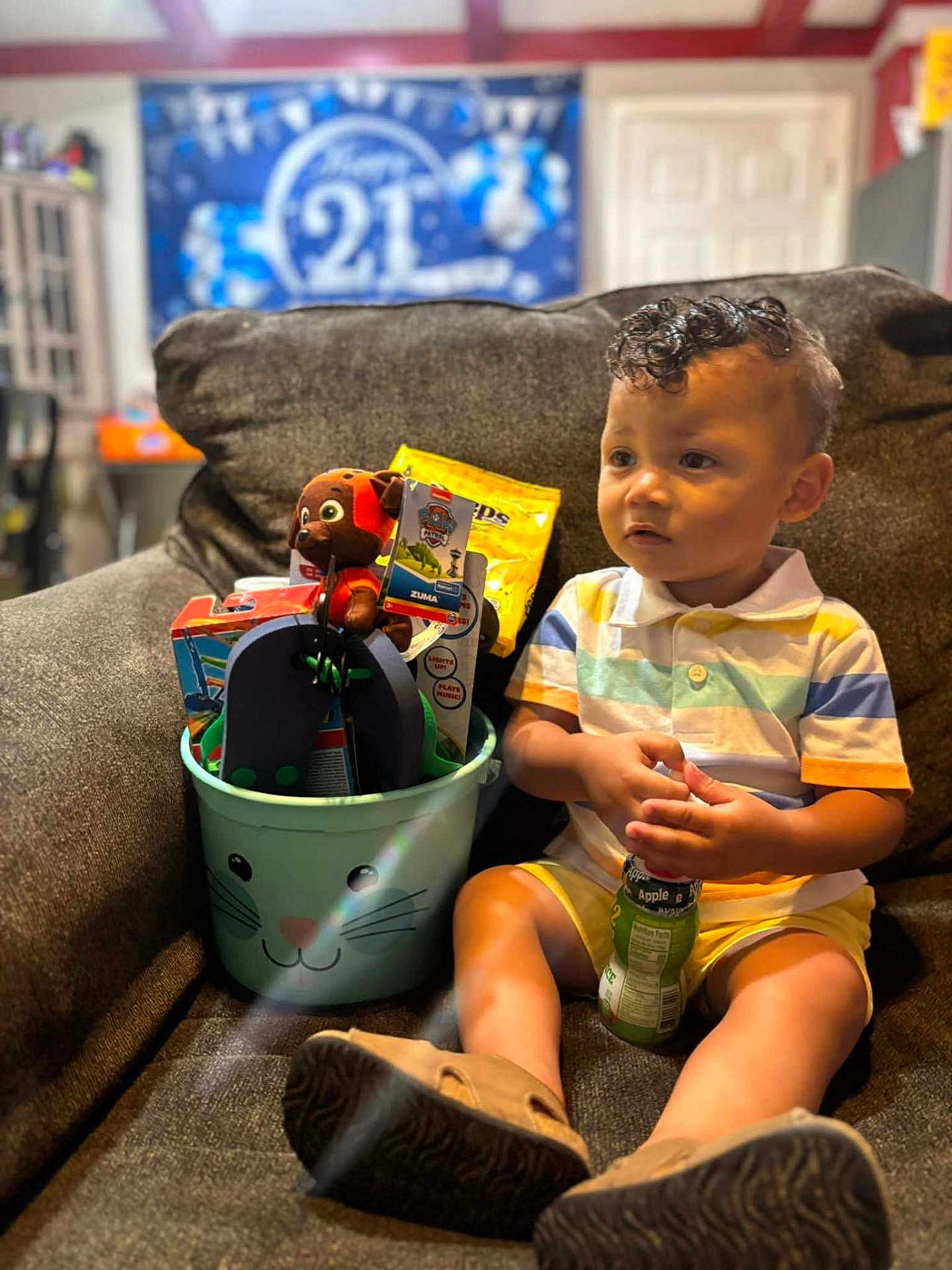 Kason is registered to the contest to win money with this photo: toddler, child, curly_hair, juice_box, toys, plush_toy, flip_flops, bucket, couch, indoor, celebration, banner, happy_21, striped_shirt, yellow_shorts, footwear, brown_couch, smiling_face, person, home