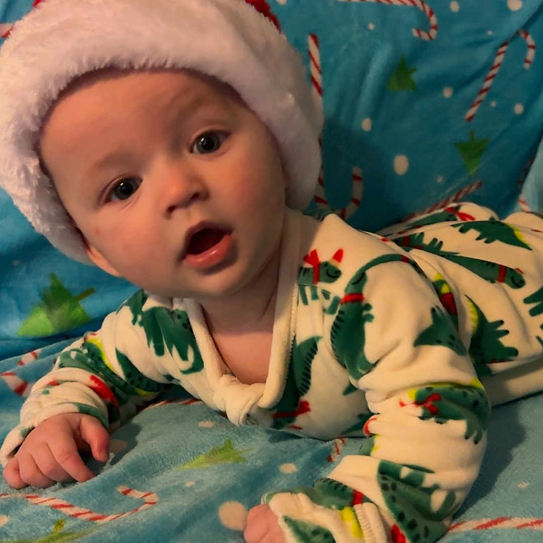 Emerson joined the competition — help win amazing prizes! baby, blanket, blue_blanket, candy_cane, child, christmas_tree, cozy, cute, dinosaur_pattern, expression, face, hands, holiday_theme, indoor, infant, lying_down, pajamas, portrait, santa_hat, soft