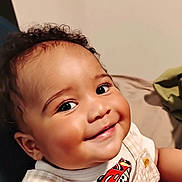 J'Khari is registered to the contest to win money with this photo: baby, child, smiling, face, curly_hair, clothing, cartoon_cars, portrait, indoor, cute, happy, infant, person, skin, toddler, cheeks, eyes, sitting, overalls, short_sleeves