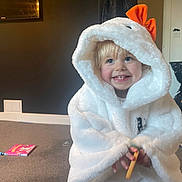 Nancy-Valentina is registered to the contest to win money with this photo: blond_hair, blue_eyes, book, carpet, child, clothing, costume, cozy, cute, happy, hood, indoor, orange_bow, person, room, small_object, smile, toddler, toy, white_fabric