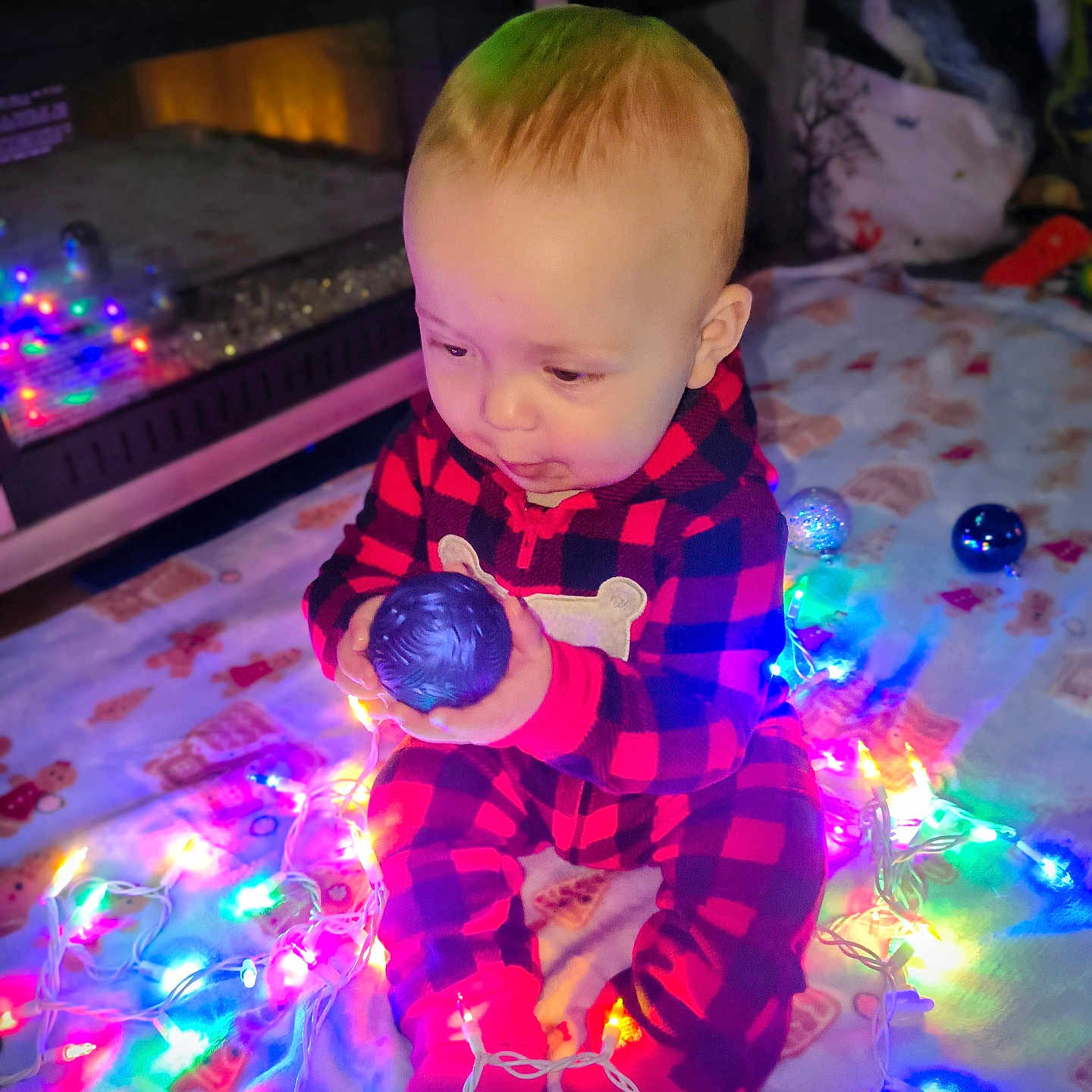 Odin joined the competition — help win amazing prizes! baby, child, christmas_lights, colorful_lights, curious, cute, decorations, festive, fireplace, floor_blanket, holiday_lights, holiday_season, indoors, infant, onesie, ornament, plaid_clothing, sitting, toy, warm_lighting