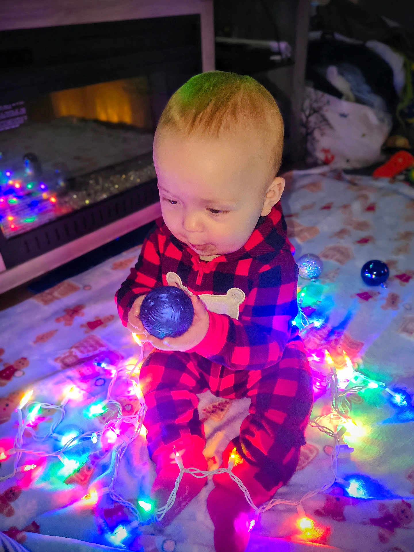 Odin joined the competition — help win amazing prizes! baby, child, infant, holiday_lights, christmas_lights, ornament, plaid_clothing, onesie, floor_blanket, fireplace, indoors, festive, colorful_lights, cute, curious, sitting, toy, warm_lighting, holiday_season, decorations