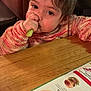 casual, child, closeup, cozy, curly_hair, expression, face, food, green_vegetable, hand, indoor, menu, person, restaurant, seated, snack, sweater, table, toddler, wooden_table