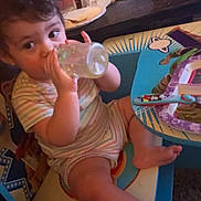 Mariana is registered to the contest to win money with this photo: bottle, casual, chair, child, curly_hair, drinking, face, food_tray, foot, hand, home, indoor, meal, person, plate, spoon, striped_clothing, table, toddler, young_child