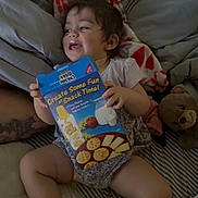 Mariana is registered to the contest to win money with this photo: toddler, child, smiling, bed, blanket, toy, teddy_bear, snack_box, cracker_box, clothing, indoor, happy, person, face, legs, hand, furniture, couch, cozy, relaxed
