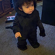 Mariana is registered to the contest to win money with this photo: toddler, child, costume, monster_costume, black_fur, indoor, carpet, snack, curly_hair, cute, hood, living_room, furniture, television, curious, standing, person, floor, toy, home
