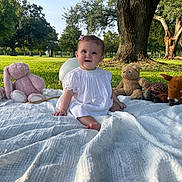 Love joined the competition — help win amazing prizes! baby, child, white_dress, blanket, plush_toys, pink_bunny, teddy_bear, turtle, fawn, grass, park, tree, sunlight, outdoor, smiling, sitting, cute, nature, greenery, daytime