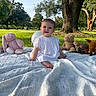 baby, child, white_dress, blanket, plush_toys, pink_bunny, teddy_bear, turtle, fawn, grass, park, tree, sunlight, outdoor, smiling, sitting, cute, nature, greenery, daytime