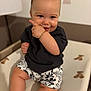 baby, child, smiling, pointing, black_shirt, floral_shorts, teddy_bear_pattern, changing_pad, indoors, cute, happy, skin, face, hand, sitting, short_hair, expression, infant, person, playful