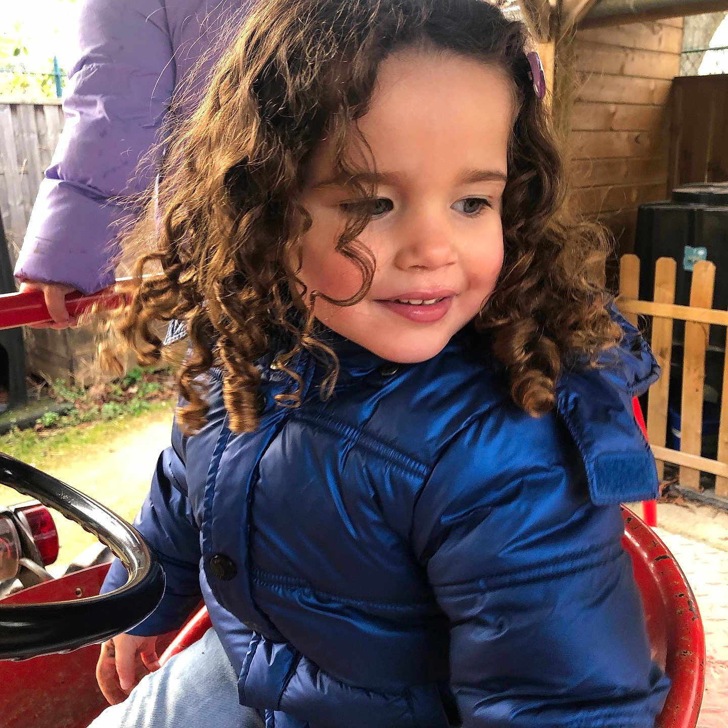 Kamila a rejoint le concours — aidez-le/la à gagner de superbes lots ! blue_jacket, child, curly_hair, daylight, face, fence, fun, happy, jeans, outdoor, person, play, playground, purple_jacket, red_toy, ride_on_toy, smile, toy, winter_clothing, wooden_shed