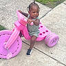 toddler, child, tricycle, pink, outdoor, sidewalk, grass, toy, play, braids, beads, sneakers, concrete, person, vehicle, summer, casual_clothing, cute, young, fun