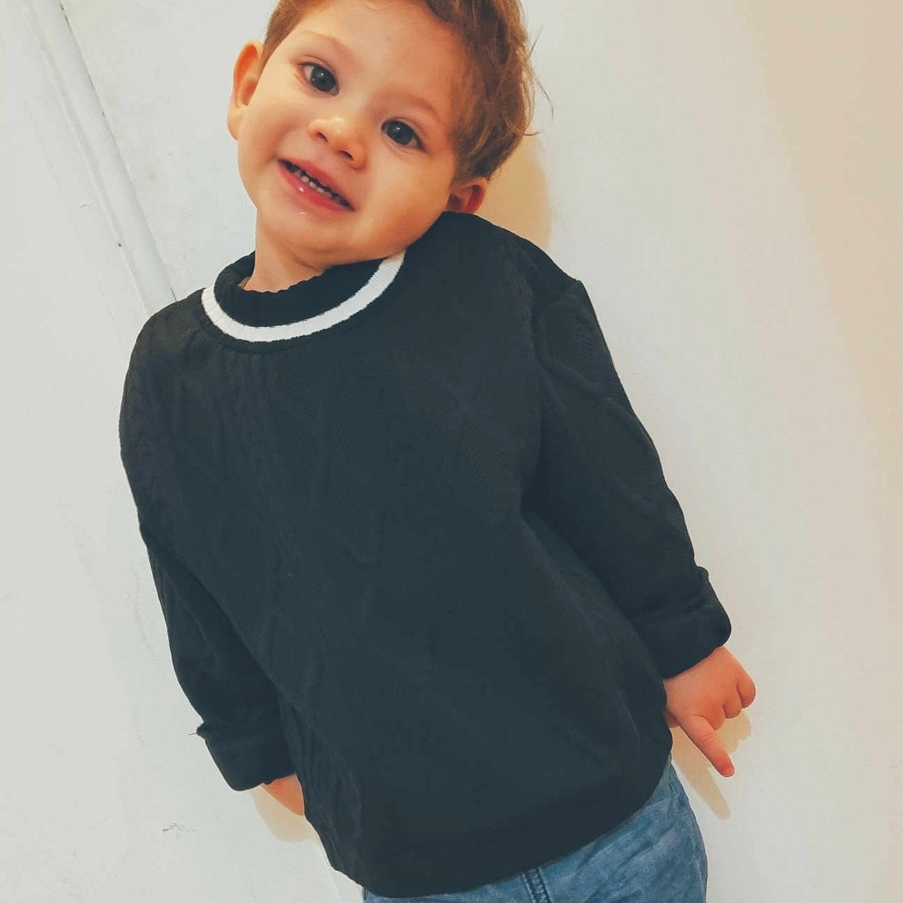 Milann participe au concours pour gagner de l'argent avec cette photo : black_sweater, blue_jeans, casual_clothing, child, cute, face, fashion, hair, happy, indoor, leaning, person, playful, portrait, posing, smiling, standing, toddler, white_wall, young_child