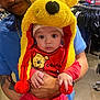 baby, child, adult, person, hat, yellow_hat, red_clothing, store, shopping, indoor, pom_pom, face, eyes, hands, holding, beard, tattoo, blue_shirt, curious, cute