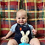 baby, child, smiling, plush_toy, bunny, overalls, striped_shirt, couch, plaid_pattern, colorful, indoor, seated, happy, cute, toy, person, face, furniture, comfort, portrait