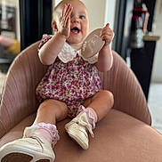 Nansi is registered to the contest to win money with this photo: baby, child, smiling, clapping, pink_chair, floral_outfit, shoes, socks, indoor, modern_interior, happy, cute, person, portrait, infant, furniture, seat, blurred_background, hands, round_sign