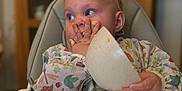 Nansi joined the competition — help win amazing prizes! baby, high_chair, food, messy, hand, flatbread, face, curious, indoor, clothing, table, meal, child, feeding, kitchen, person, cute, expression, sitting, infant