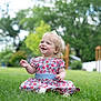 toddler, child, grass, outdoor, smiling, dress, flower_pattern, greenery, backyard, nature, happy, person, sitting, young_child, sunlight, daytime, cute, casual, playful, blond_hair