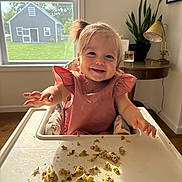 Piper is registered to the contest to win money with this photo: toddler, child, high_chair, messy_food, smiling, indoor, window, natural_light, pink_dress, hair_tied, plant, table_lamp, wooden_floor, family_photo, home_interior, happy, baby, face, food, cute