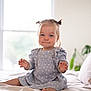 child, toddler, baby, pigtails, blue_dress, polka_dot, bed, pillow, window, natural_light, indoor, smile, sitting, cute, hair, furniture, plant, soft_light, portrait, happy