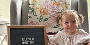 Piper is registered to the contest to win money with this photo: baby, child, infant, armchair, blanket, plush_toy, sign, window, curtains, pigtails, smiling, indoors, milestone, clothing, feet, face, person, decor, natural_light, happy