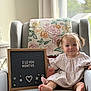 baby, child, infant, armchair, blanket, plush_toy, sign, window, curtains, pigtails, smiling, indoors, milestone, clothing, feet, face, person, decor, natural_light, happy