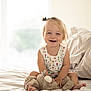 toddler, child, smiling, bed, stuffed_animal, plush_toy, indoor, natural_light, pony_tail, happy, cute, person, clothing, white_bedspread, soft_toys, infant, sitting, cozy, home, portrait