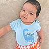 adorable, baby, blue_shirt, child, clothing, cute, dinosaur_print, eyes, face, hair, indoors, infant, lying_down, orange_pants, patterned_clothing, person, portrait, skin, smile, soft_surface
