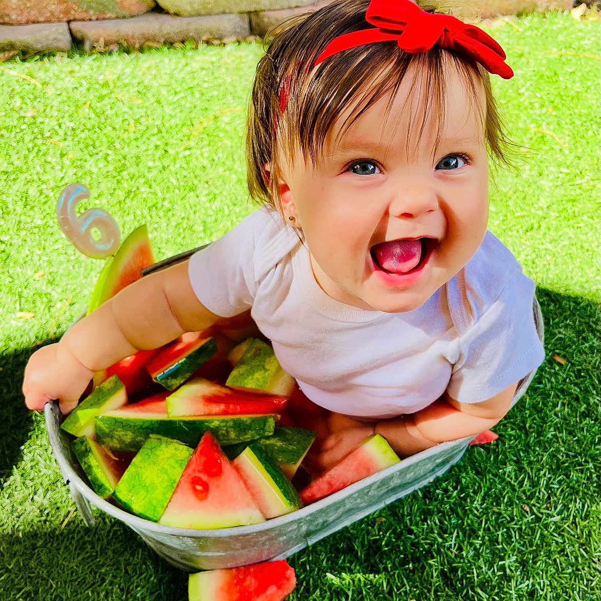 Gia joined the competition — help win amazing prizes! baby, bucket, cheerful, child, cute, fruit, grass, green, happy, headband, nature, outdoor, playful, red_bow, smiling, summer, sunlight, toddler, watermelon, white_shirt