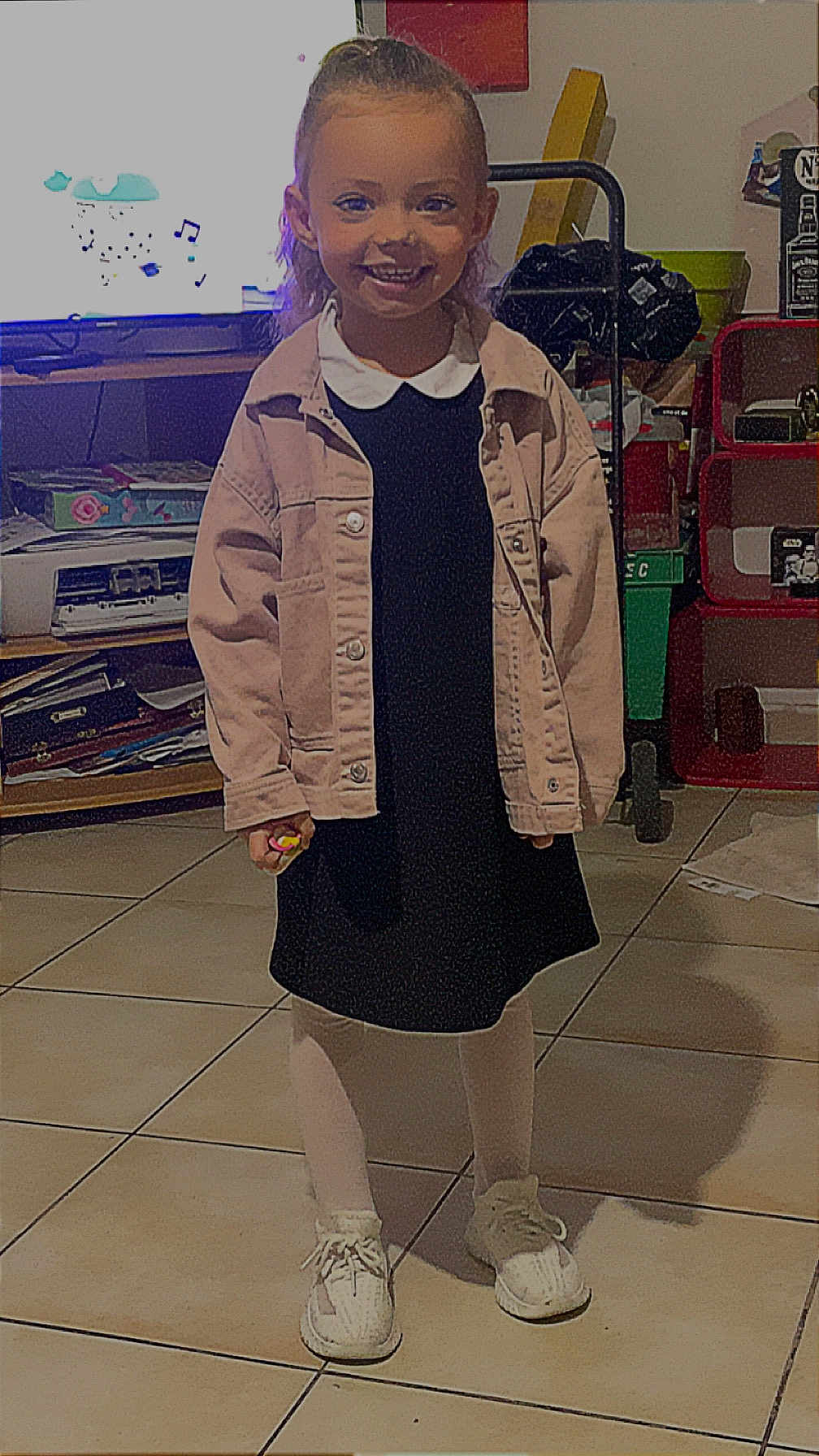 Gabryelle participe au concours pour gagner de l'argent avec cette photo : child, smiling, indoor, tiled_floor, jacket, dress, tights, sneakers, person, furniture, television, shelf, clutter, toy, happy, standing, floor, casual_clothing, hair, young