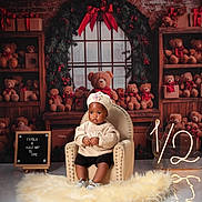 Fayola is registered to the contest to win money with this photo: baby, child, chair, teddy_bear, holiday_decor, gift_box, window, rug, knit_hat, sweater, floor, signboard, light_number, red_bow, wooden_shelves, plush_toy, indoor, portrait, celebration, smiling