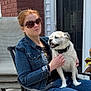 Sitich is registered to the contest to win money with this photo: woman, dog, sunglasses, denim_jacket, chair, porch, brick_wall, white_door, black_lantern, casual_clothing, pet, sitting, outdoor, daylight, smiling_dog, relaxed, hand_holding, jeans, plant, metal_chair