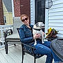 Sitich is registered to the contest to win money with this photo: woman, dog, chair, patio, outdoor, sunglasses, denim_jacket, brick_chimney, white_siding, potted_plant, table, casual_clothing, smiling, pet, relaxing, daytime, home, fence, footwear, jeans