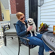Sitich joined the competition — help win amazing prizes! woman, dog, sunglasses, denim_jacket, jeans, black_shirt, patio, chair, table, potted_plant, brick_wall, door, can, outdoor, smiling_dog, casual, relaxed, daylight, footwear, happy