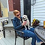 woman, dog, sunglasses, denim_jacket, jeans, black_shirt, patio, chair, table, potted_plant, brick_wall, door, can, outdoor, smiling_dog, casual, relaxed, daylight, footwear, happy