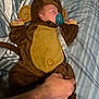 baby, sleeping, monkey_costume, pacifier, bed, striped_bedspread, hand, costume, child, cute, infant, soft_toy, brown, yellow, clothing, resting, indoors, cozy, person, pacifier_clip